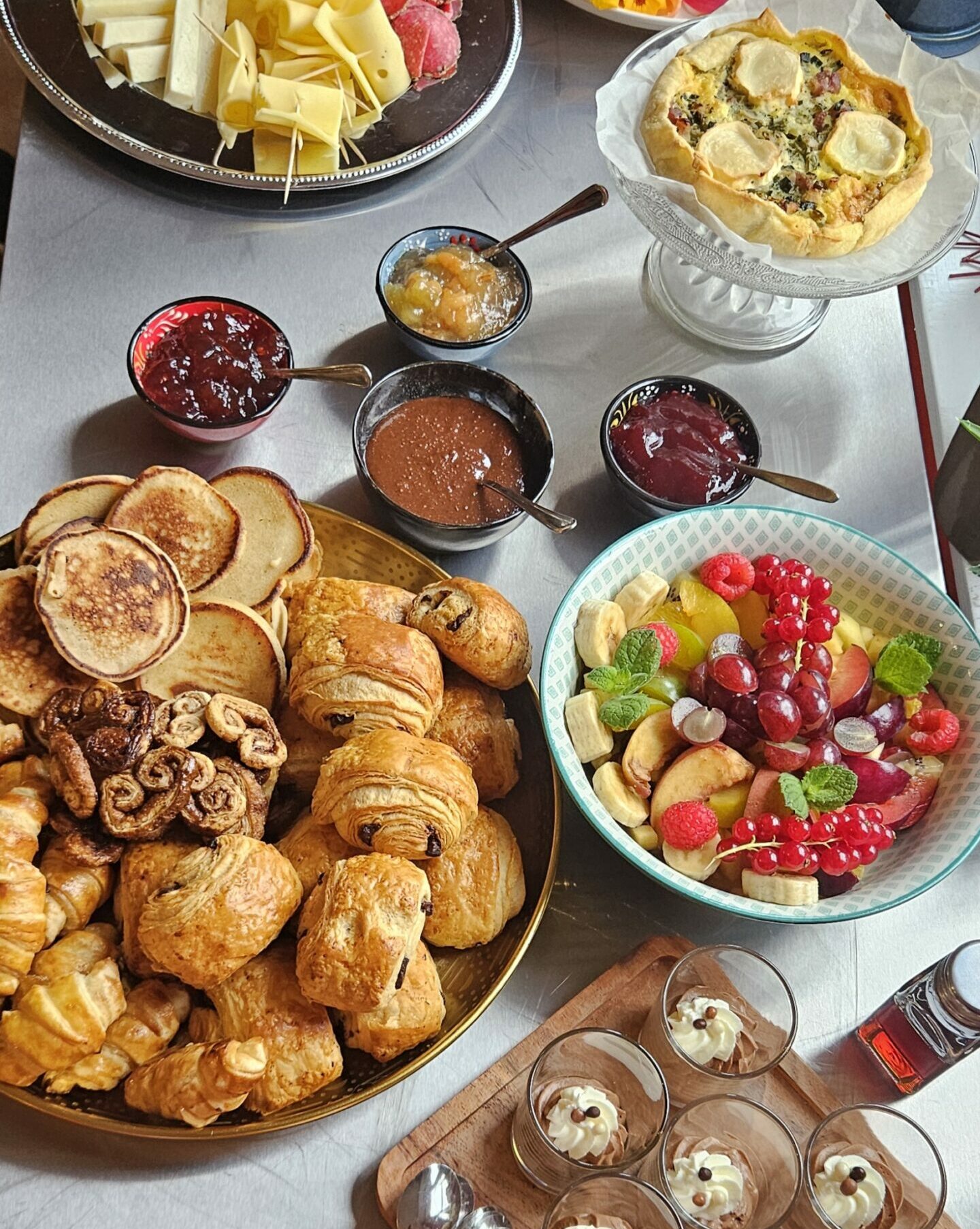Buffet brunch gourmand Simple & Bon composé de pains maison, viennoiseries, fromages, charcuteries, saumon fumé, quiches, omelette et lard, confitures maison, pancakes, salade de fruits de saison et verrines sucrées. Présentation colorée et conviviale sur table.