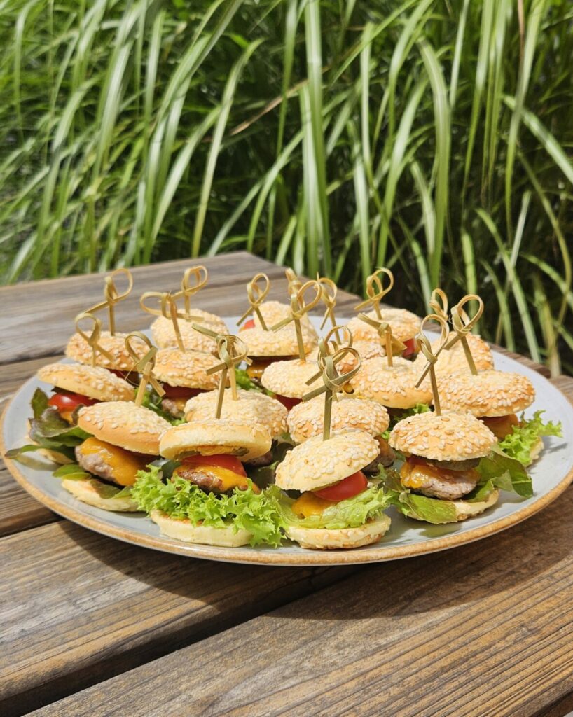 Plateau de mini burgers apéritifs garnis de salade, fromage et tomate, fixés avec un pic en bois sur des pains briochés au sésame.