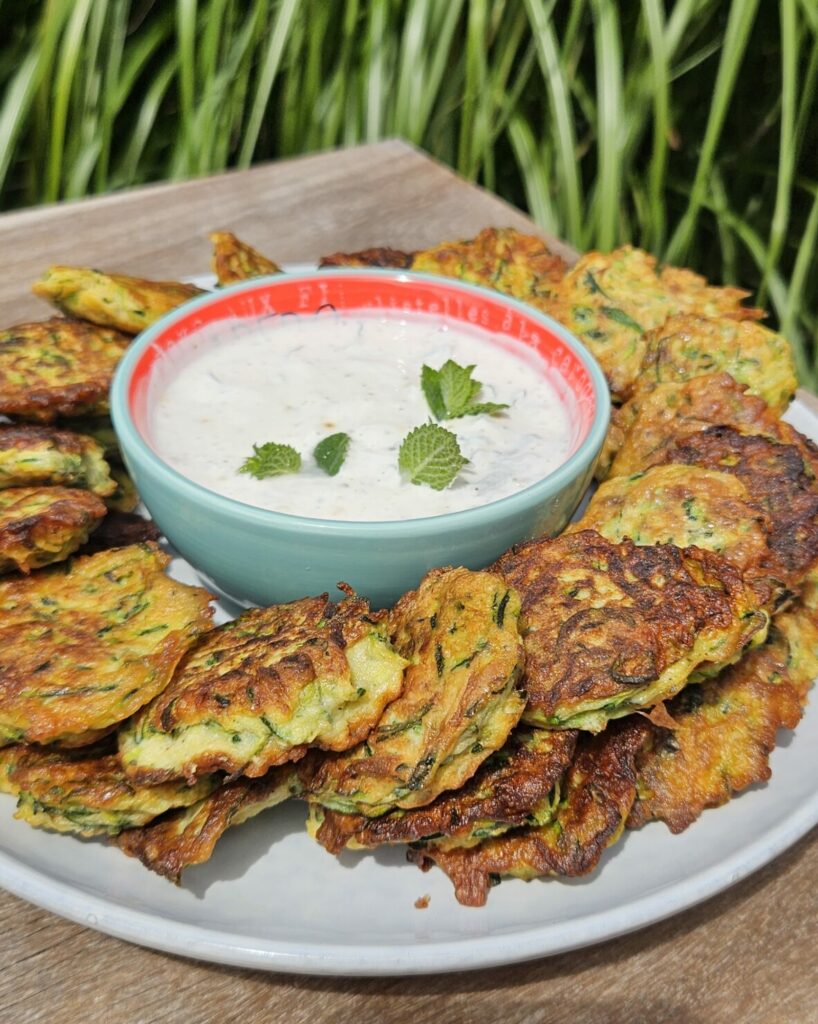 Assiette de rösti de courgette dorés et croustillants, accompagnés d’une sauce crémeuse à l’ail décorée de feuilles de menthe fraîche.