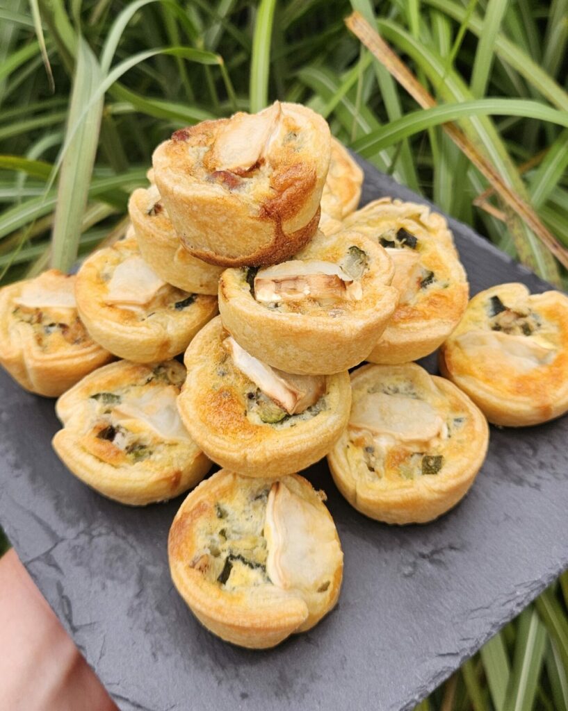 Assortiment de mini quiches dorées garnies de légumes et fromage, présentées sur une ardoise.