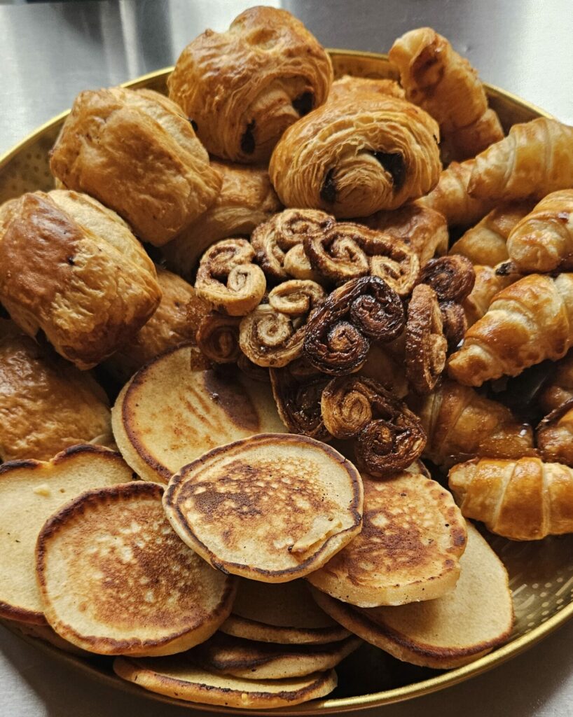 Assortiment de viennoiseries maison avec croissants, pains au chocolat, palmiers et pancakes, présenté sur un grand plateau doré.