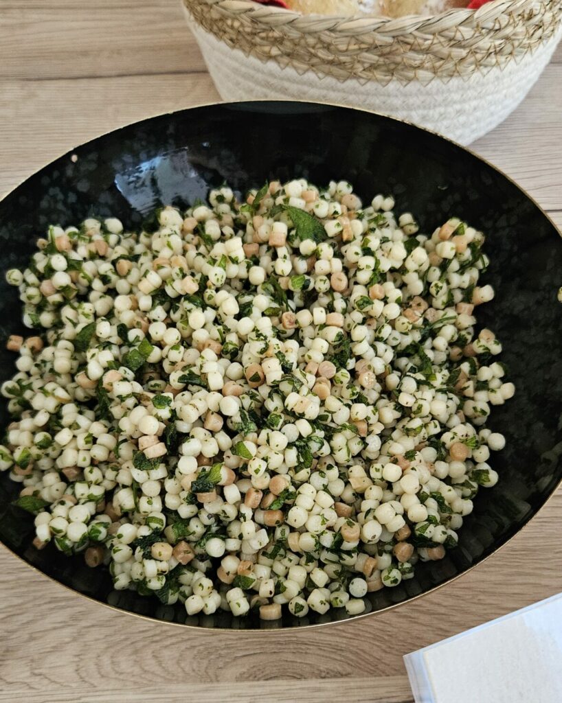 Bol noir de salade de fregola sarda aux herbes fraîches et persil, avec une corbeille de petits pains en arrière-plan.