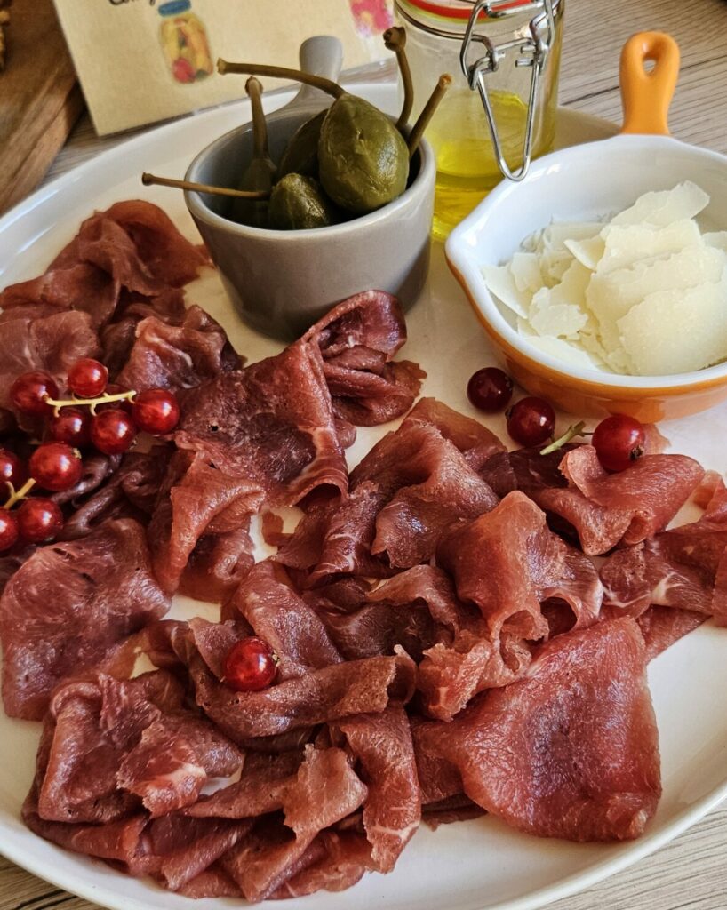 Assiette d’antipasti italiens composée de viande séchée en tranches fines, copeaux de parmesan, câpres, huile d’olive et groseilles rouges en décoration.
