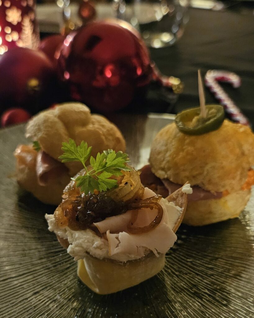 Trio d’amuses-bouches festifs sur assiette noire, comprenant chou à la crème salé, mini burger et bouchée garnie de volaille et d’oignons caramélisés, décor de table de Noël en fond.