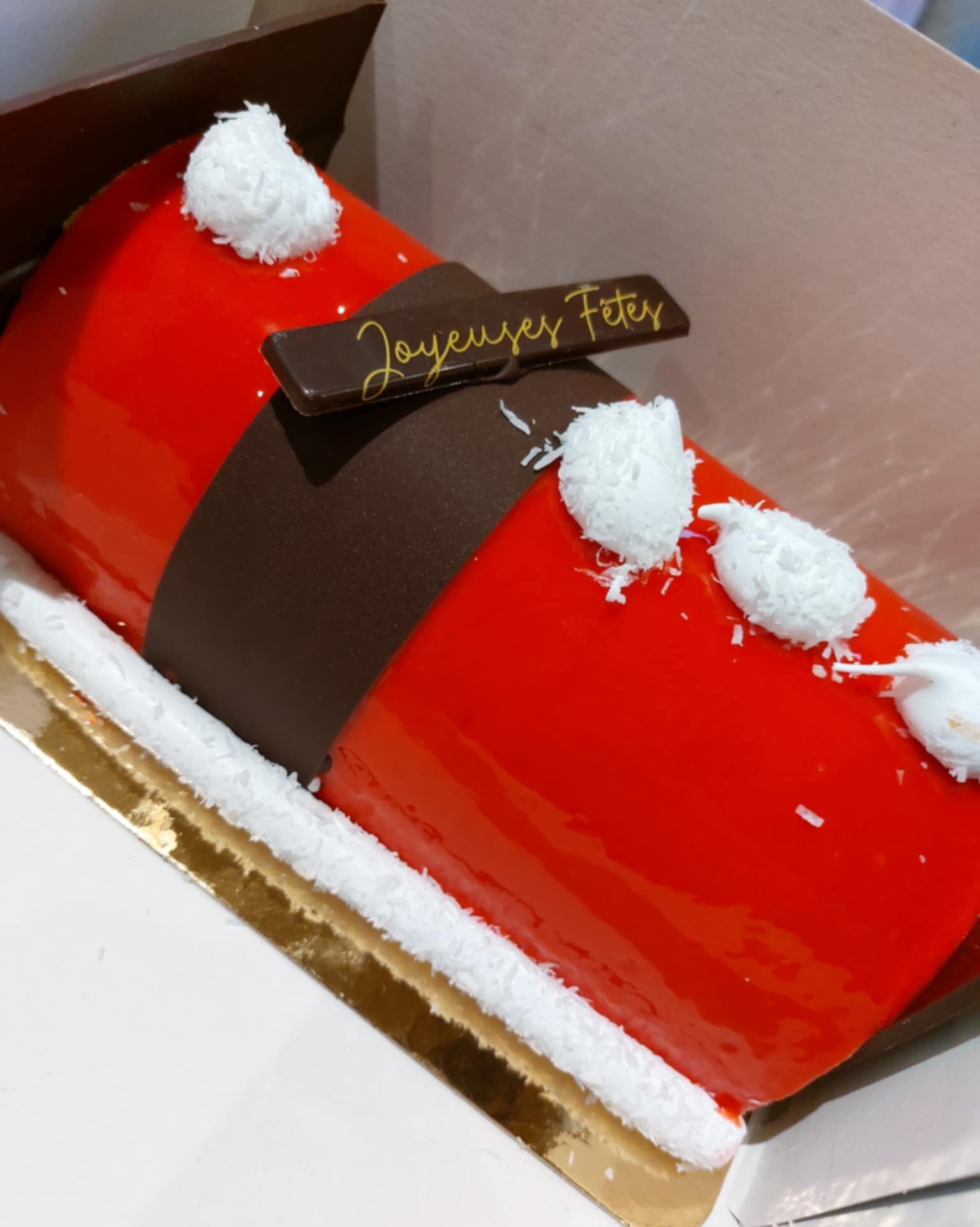 Bûche de Noël rouge brillante façon manteau du Père Noël, décorée de chocolat noir et de flocons de coco, avec l’inscription “Joyeuses Fêtes”.