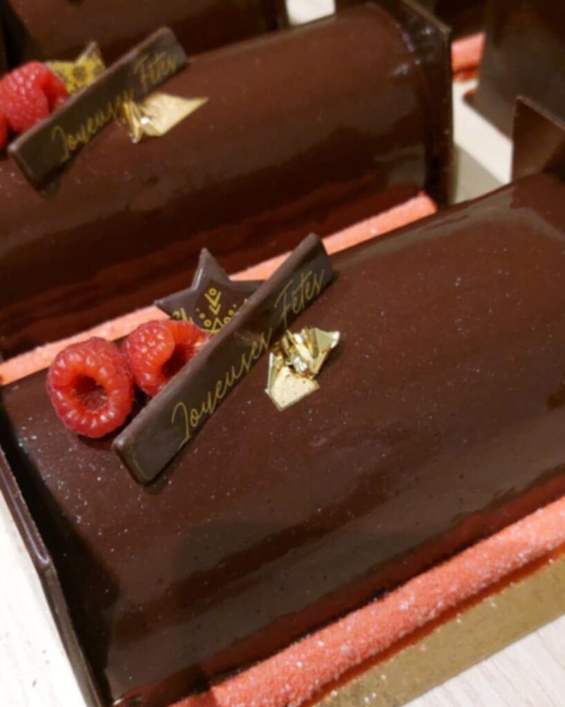 Bûches de Noël au chocolat noir brillant décorées de framboises fraîches, d’éclats dorés et d’une plaque “Joyeuses Fêtes”.