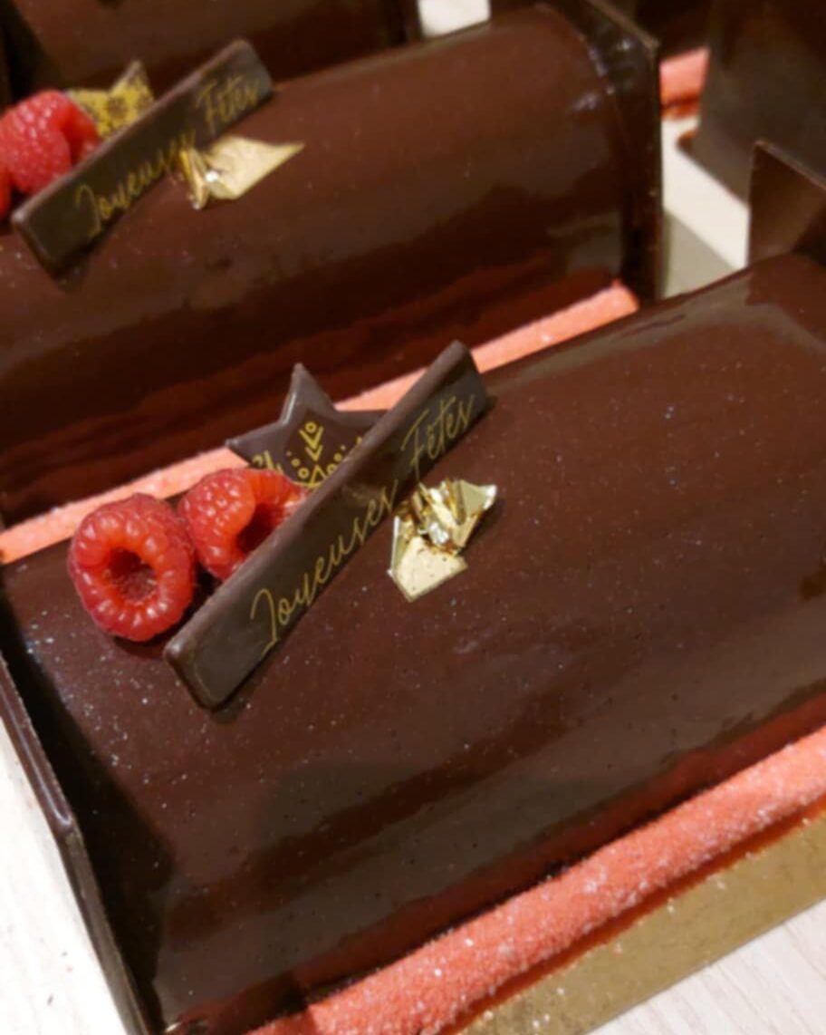 Bûches de Noël au chocolat noir brillant décorées de framboises fraîches, d’éclats dorés et d’une plaque “Joyeuses Fêtes”.