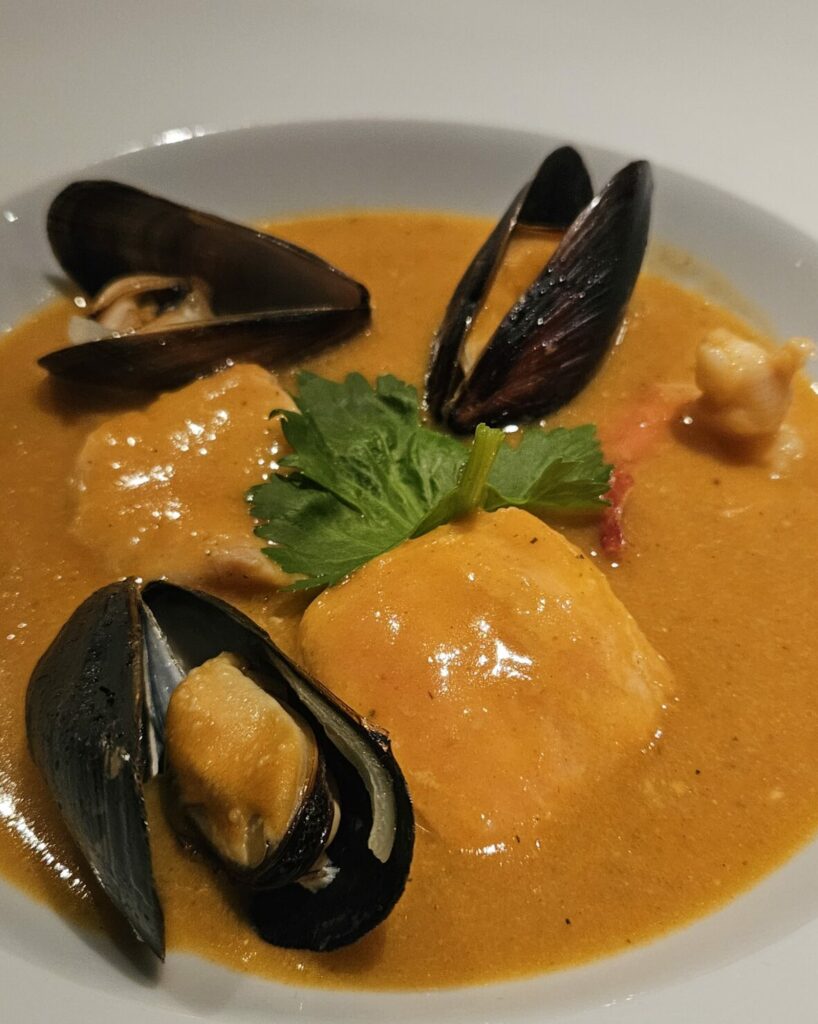 ssiette de soupe de poisson maison garnie de moules, morceaux de poisson et croûtons, servie dans une sauce onctueuse orangée aux saveurs iodées.