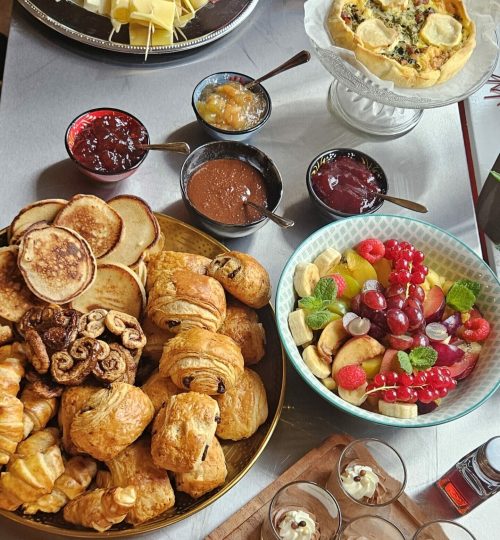 Buffet brunch gourmand Simple & Bon composé de pains maison, viennoiseries, fromages, charcuteries, saumon fumé, quiches, omelette et lard, confitures maison, pancakes, salade de fruits de saison et verrines sucrées. Présentation colorée et conviviale sur table.